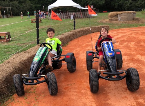 Two children smiling while sitting in go-karts on a dirt track, with hay bales and a festive background.