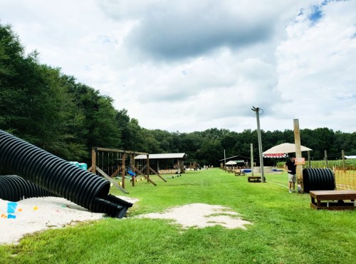 A playground with slides and tunnels on a grassy field, surrounded by trees under a cloudy sky.