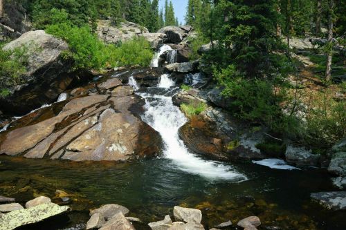 A serene waterfall cascades over rocky terrain, surrounded by lush greenery and tall trees.