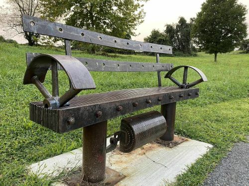 A unique metal bench with industrial design, featuring curved armrests and a textured seat, set in a grassy area.