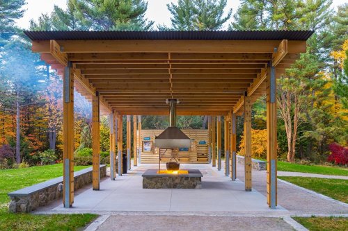 A modern pavilion with a stone fire pit, surrounded by trees and colorful autumn foliage.