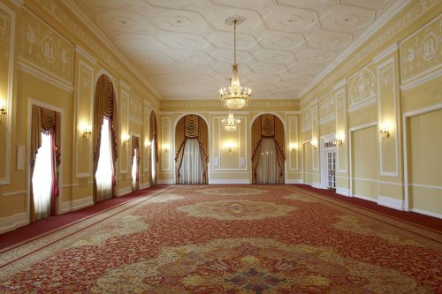 A spacious, elegantly decorated room with ornate ceilings, chandeliers, and rich carpet, featuring large windows and drapes.