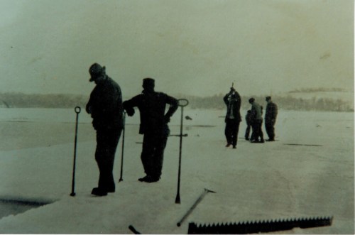 Workers on a snowy landscape, using tools to cut or manage ice, with a distant view of trees and hills.