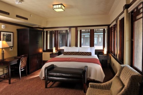 Cozy hotel room featuring a king bed, desk, armchair, and large windows with natural light. Warm, inviting decor.