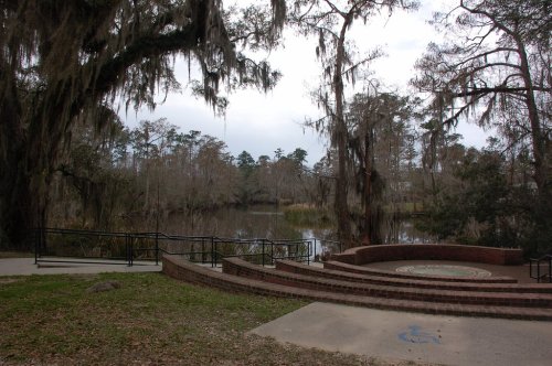 A serene view of a lake surrounded by trees draped in Spanish moss, with a brick seating area in the foreground.
