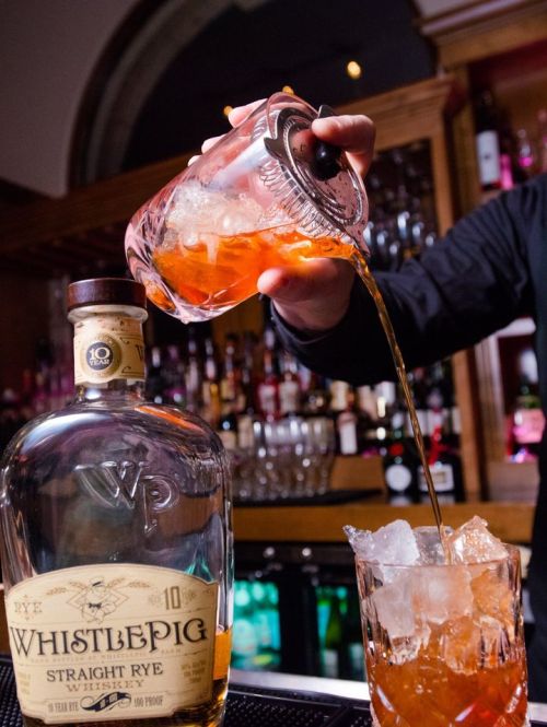 A bartender pours whiskey from a bottle into a glass filled with ice, with a bar backdrop.