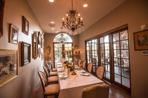 Elegant dining room with a long table, chandelier, and framed artwork on the walls, leading to a glass door.