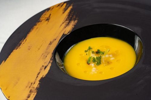 A vibrant yellow soup served in a black plate with a decorative gold brushstroke, garnished with herbs and diced onions.