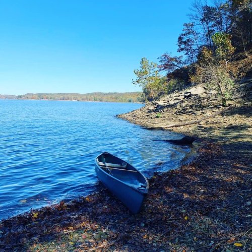 A canoe rests on a rocky shore by a calm lake, surrounded by trees and a clear blue sky.