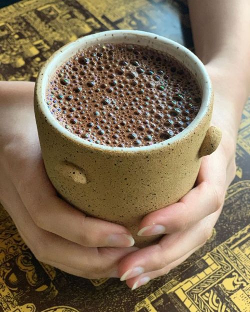 A person holds a textured ceramic mug filled with frothy chocolate drink, resting on a patterned table.