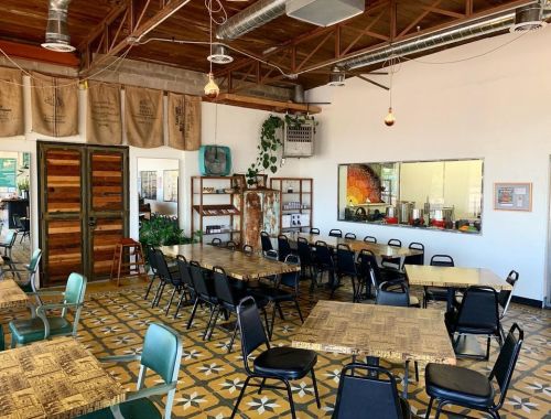 A spacious, modern restaurant interior with wooden tables, black chairs, and decorative plants.