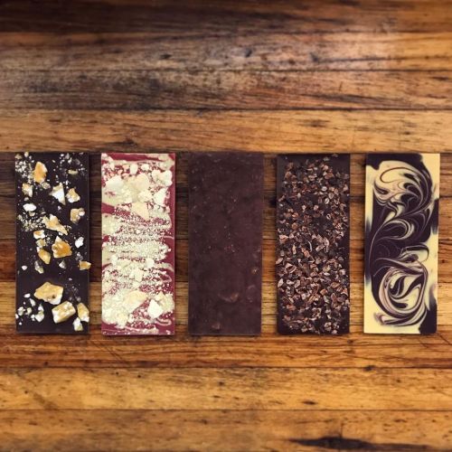 Five chocolate bars with various toppings, including nuts, swirls, and crushed cocoa, arranged on a wooden surface.