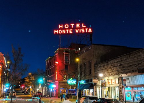 Neon sign for Hotel Monte Vista illuminated at night, with a lively street scene and buildings in the foreground.