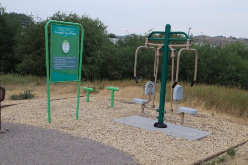 Outdoor fitness area featuring a seated chest press machine and informational sign, surrounded by gravel and grass.
