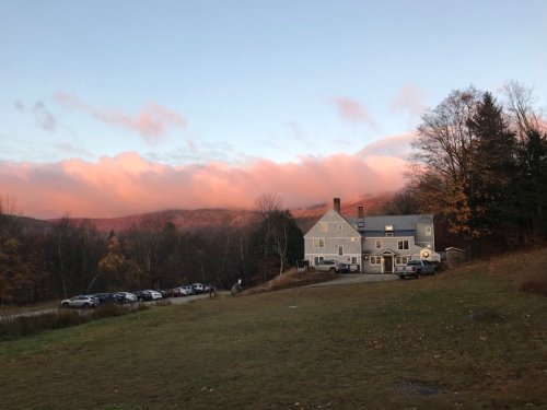 A gray house surrounded by trees and parked cars, with a colorful sunset over the mountains in the background.