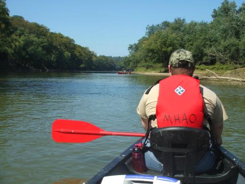A person in a red life jacket paddles a canoe on a calm river surrounded by trees under a clear blue sky.