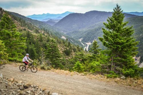 A cyclist rides on a dirt path overlooking a mountainous landscape with trees and a winding river below.
