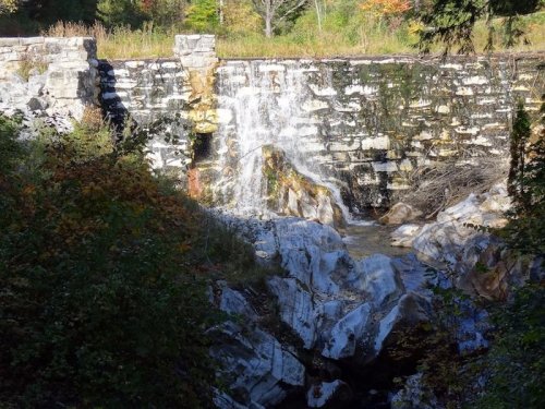 A weathered stone dam with a small waterfall, surrounded by lush greenery and rocky terrain.