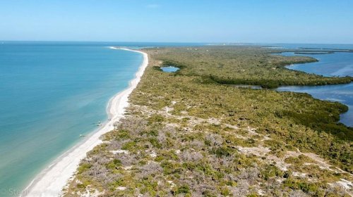 Aerial view of a sandy coastline bordered by lush greenery and calm blue waters under a clear sky.