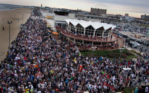 A large crowd gathers along a beachside boardwalk, with a restaurant and ocean in the background.