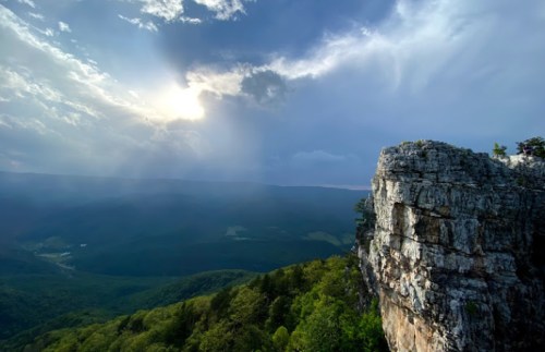 A dramatic landscape featuring a rocky cliff overlooking lush green valleys and a cloudy sky with sunlight breaking through.