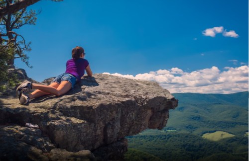 A person lies on a rocky ledge, gazing at a scenic view of mountains and blue skies with fluffy clouds.