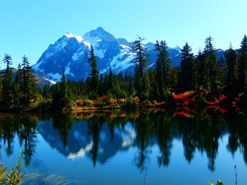 A serene mountain landscape with snow-capped peaks, lush green trees, and colorful autumn foliage reflected in a calm lake.