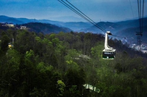 A green gondola glides over lush, green mountains under a cloudy sky, with distant hills visible in the background.