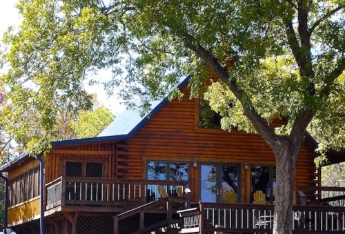 A wooden cabin with a blue roof, surrounded by trees and featuring a large deck.