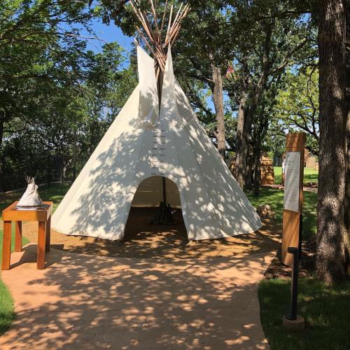 A white teepee stands among trees, with a wooden table nearby and informational signage in a sunny outdoor setting.