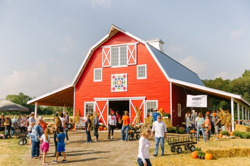 A vibrant red barn with a quilt pattern, surrounded by people, pumpkins, and hay bales on a sunny day.