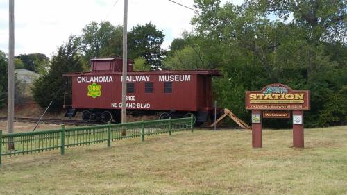 A red train caboose labeled "Oklahoma Railway Museum" beside a sign for the museum, set in a grassy area with trees.
