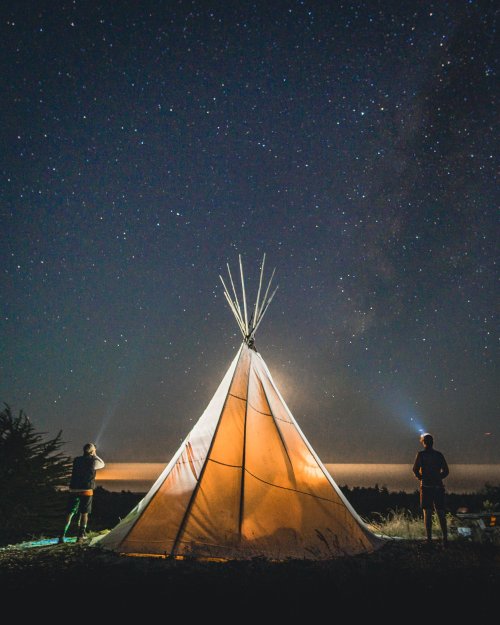 Two people stand by a lit teepee under a starry night sky, holding flashlights and enjoying the serene atmosphere.