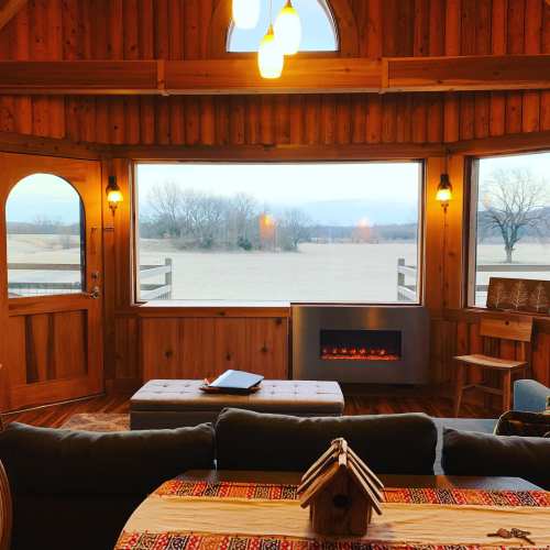 Cozy wooden interior with a view of a field, featuring a fireplace, a coffee table, and warm lighting.