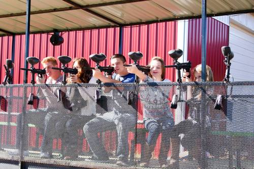 A group of five young people sitting on a platform, aiming paintball guns at a target.