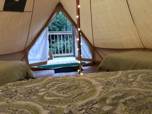 Interior of a tent with two beds, a patterned blanket, and a view of a deck and trees through the open entrance.