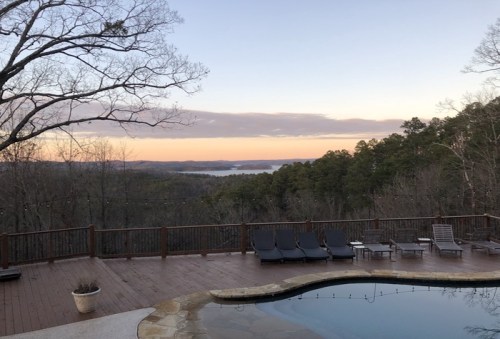 A serene view of a poolside deck overlooking a misty valley and trees at sunrise.