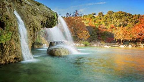 A serene waterfall cascades over rocks into a tranquil pool, surrounded by vibrant autumn foliage and clear blue skies.