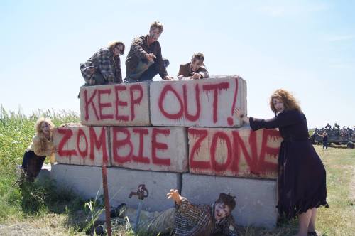 A group of people in tattered clothes poses by a concrete barrier with "KEEP OUT! ZOMBIE ZONE" painted in red.