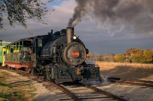 A vintage steam locomotive with smoke billowing, pulling a green passenger car along a scenic railway track.
