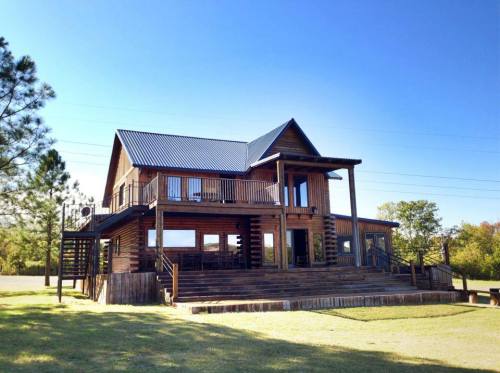 A large wooden cabin with a metal roof, featuring a spacious deck and surrounded by trees under a clear blue sky.