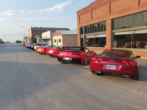 A row of red and white Corvettes parked along a street with brick buildings in the background.