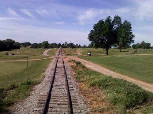 A view of a grassy landscape with railway tracks leading into the distance, flanked by paths and trees.