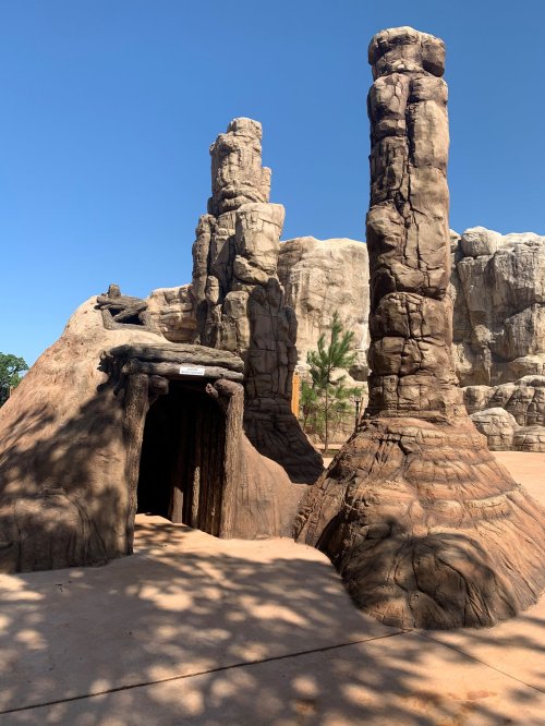 Rock formations resembling towers stand beside a cave entrance, set against a clear blue sky.