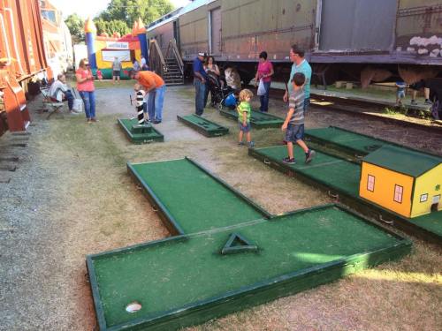 A mini-golf setup with colorful obstacles, children and adults playing near vintage train cars in a grassy area.