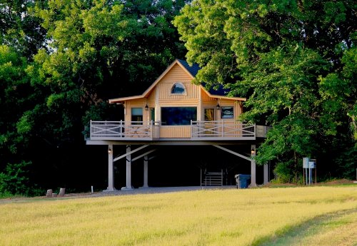 A charming yellow house on stilts surrounded by lush green trees and a grassy area.