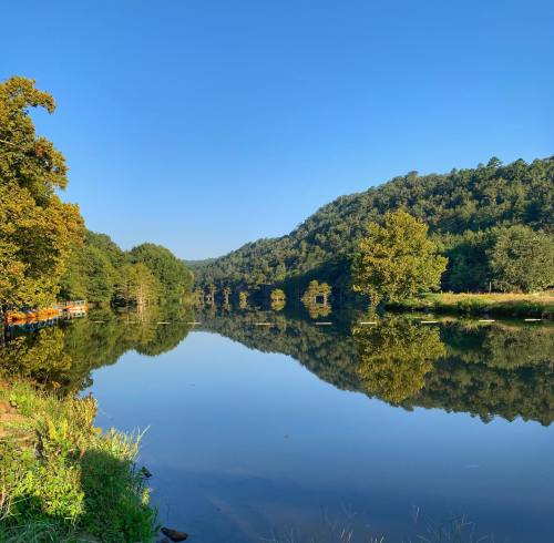 A serene lake reflecting trees and blue sky, surrounded by lush green hills on a clear day.