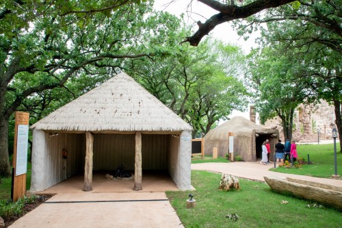 Two traditional-style huts with thatched roofs in a green park, surrounded by trees and visitors exploring the area.