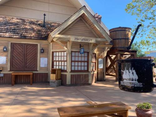 A rustic building labeled "Prosperity" with wooden benches and a water tower nearby, under a clear blue sky.