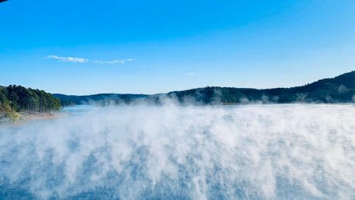 Mist rises over a serene lake surrounded by green hills under a clear blue sky.